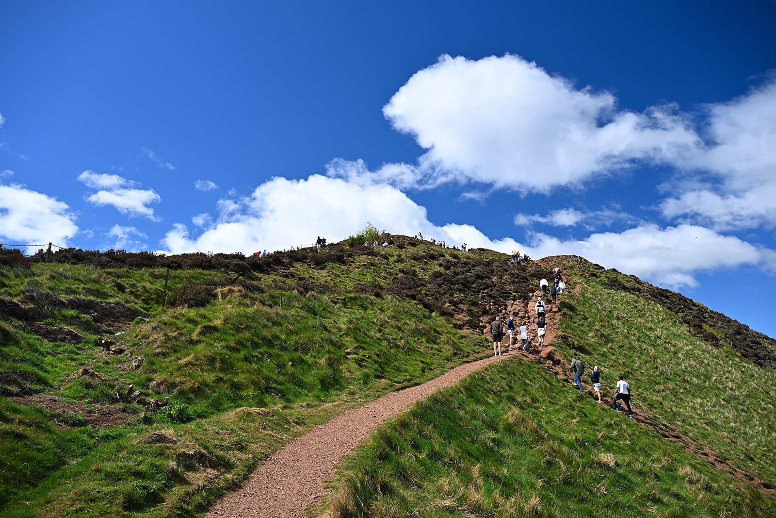Walkers climbing the grassy path towards the summit of Arthur’s Seat under a blue sky