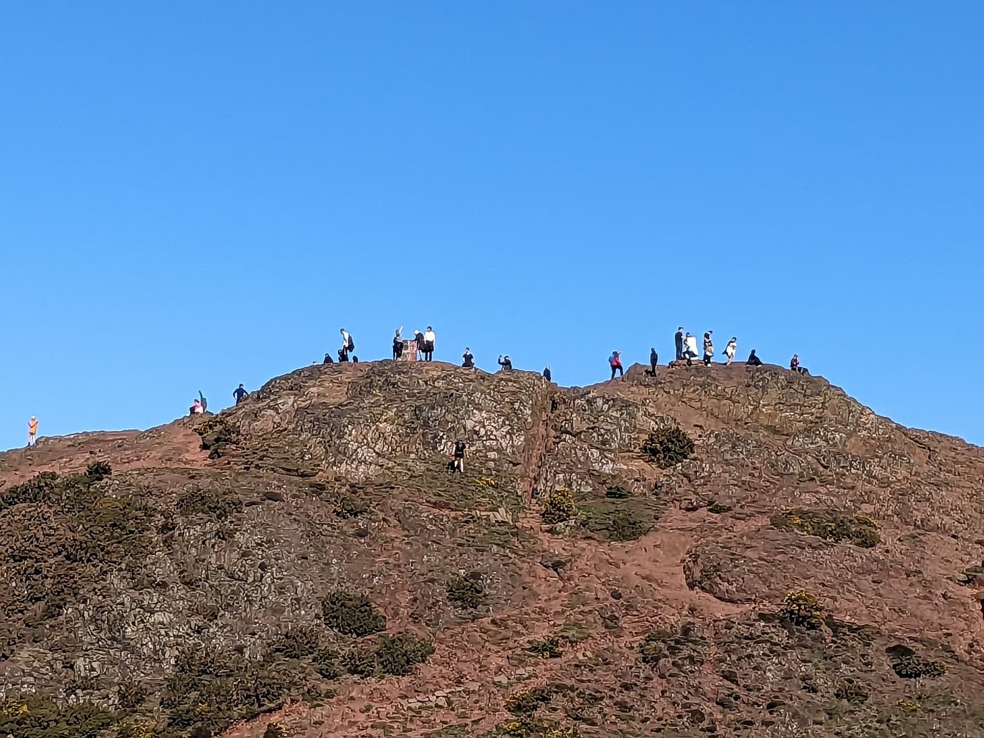 People standing on the bare rocky summit of Arthur’s Seat against a clear blue sky